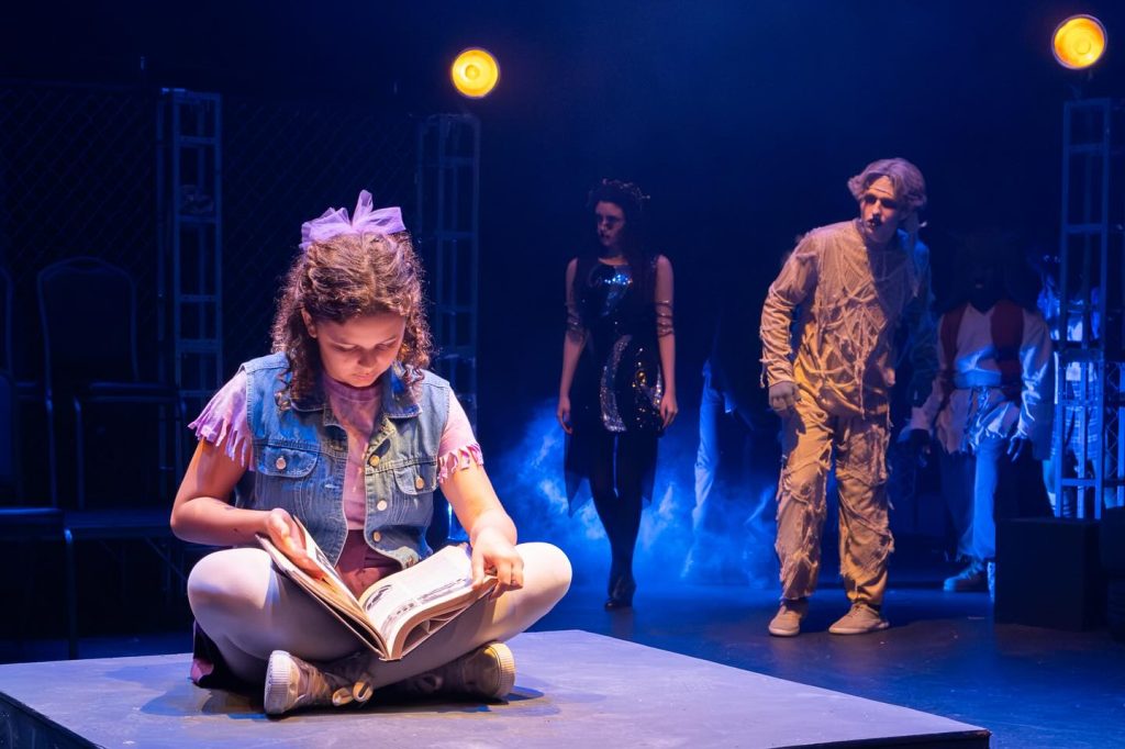 little girl reading a book onstage with monsters behind her 