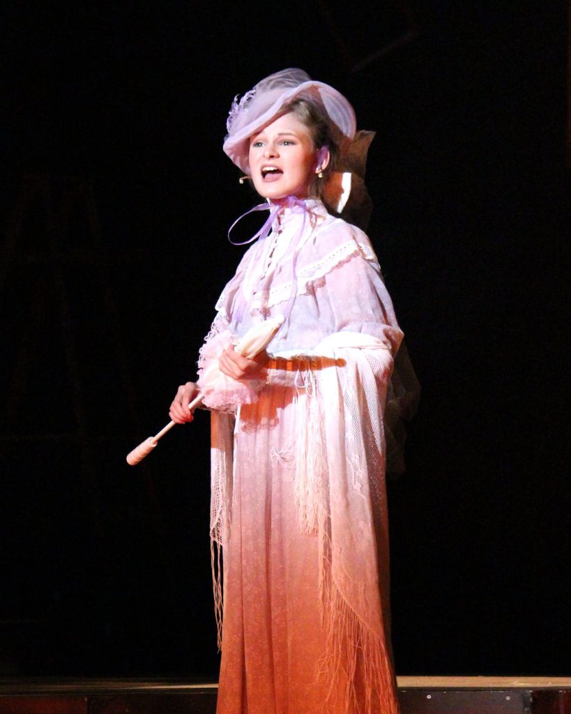 Actor with pink dress, hat and parasol singing 