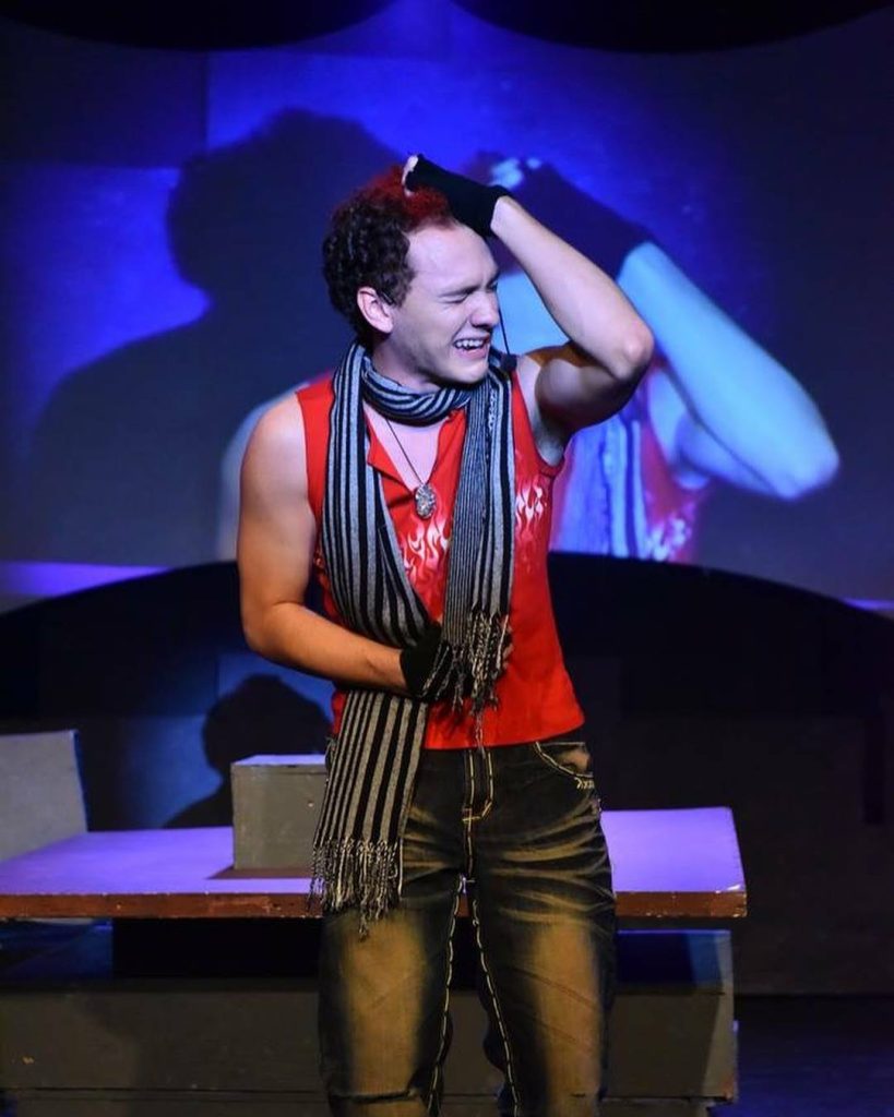 Actor performing in red cut off shirt with scarf, emotional singing Judas 